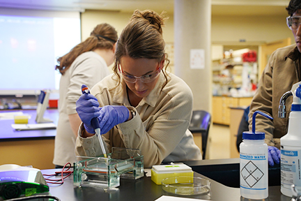 A WVC biology student loads gel with a pipette during a gel electrophoresis lab, while another student looks on.