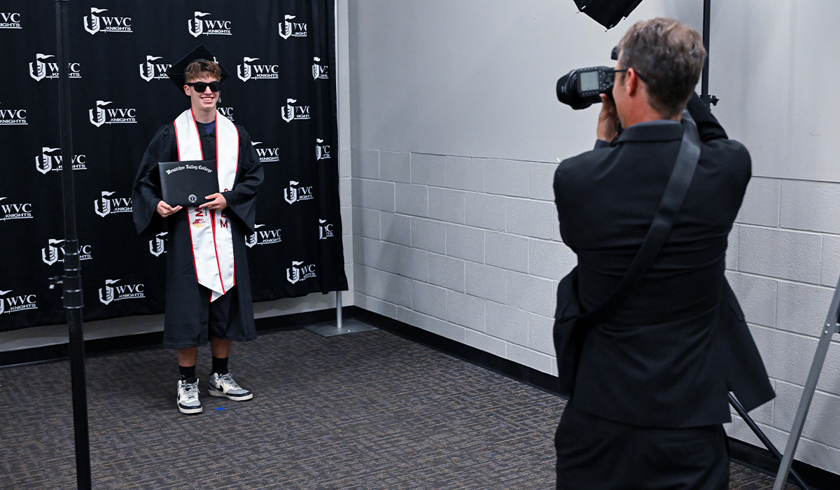 A graduate standing against a WVC Knights backdrop and holding his diploma prepares to have his photo taken by a professional photographer.
