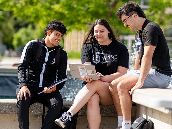Three students wearing black and white Knights gear sit at the campus fountain and look at a laptop.