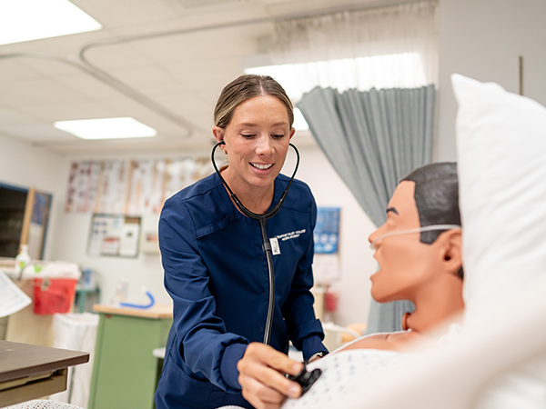 A WVC Omak nursing student wearing a stethoscope and blue scrubs checks the pulse on a manikin.