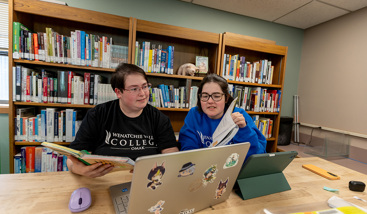 Two students sit at a table in front of a laptop and look at a book together.