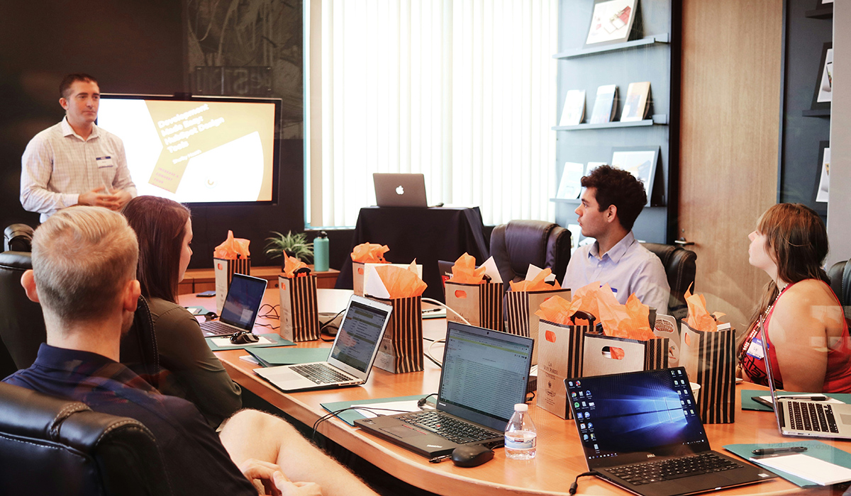 A group of marketing employees sit around a table with their laptops and listen to a presenter.