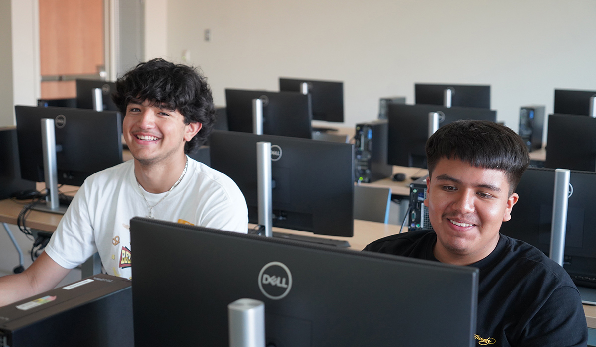 Two students sit in a computer lab and work on computers.
