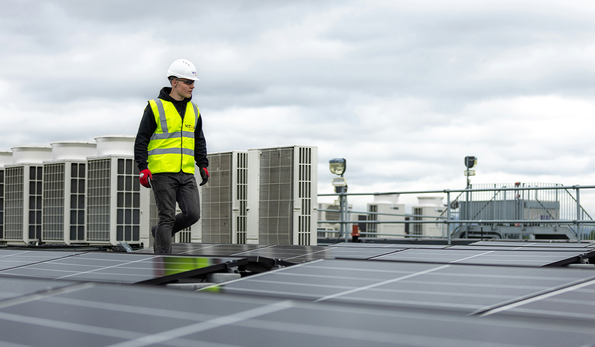 An HVAC technician walks alongside a row of heat pumps on a roof.