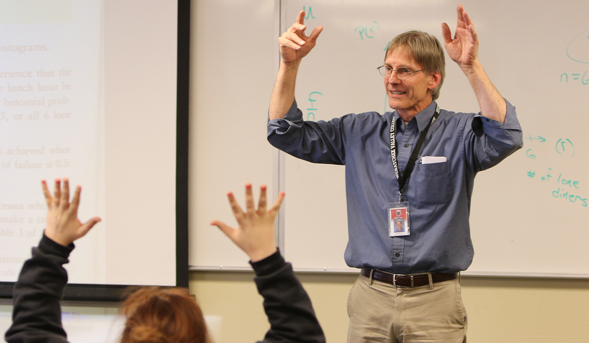 A math instructor and his student both hold their hands up in the air in a classroom.