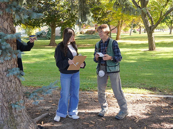 A biologist talks with a student who is holding a clipboard while they stand among a grove of trees.