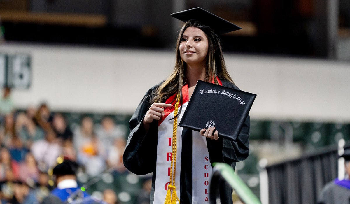 A TRIO graduate standing on the stage during the commencement ceremony points to her diploma cover.